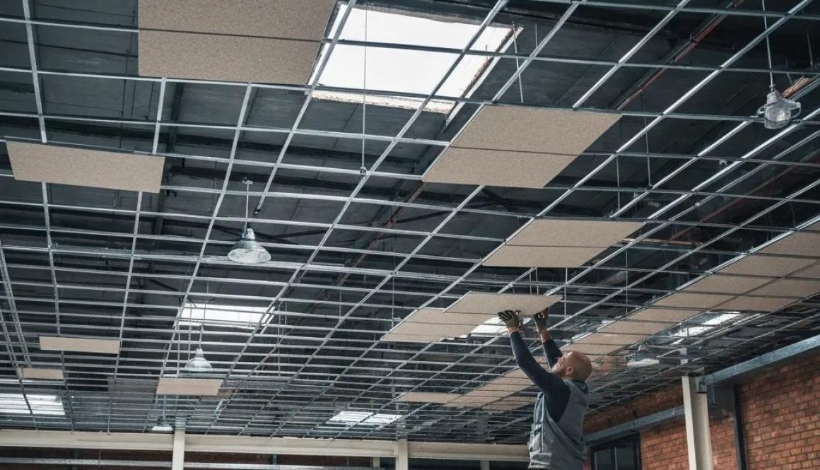 Worker installing suspended ceiling tiles in a commercial office space, showcasing modular ceiling system and overhead lighting.